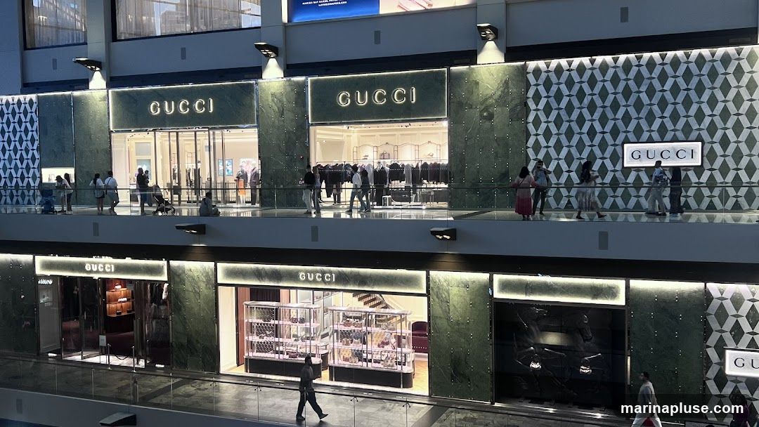 A multi-level Gucci store with prominent signage on both levels. The upper level features a large illuminated "GUCCI" sign above a glass entrance, with mannequins displaying clothing visible inside. The facade is decorated with green marble and a geometric pattern. The lower level also displays "GUCCI" signage and has large display windows showcasing handbags and accessories. People are seen browsing on both levels.