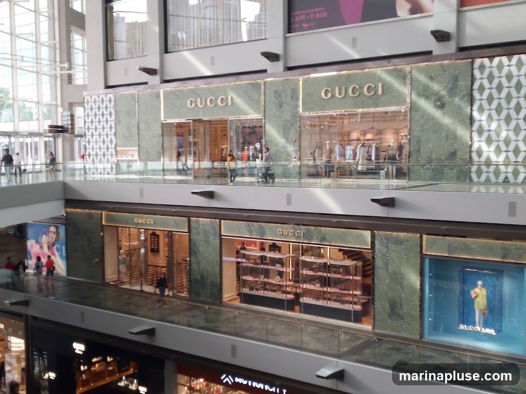 Multiple Gucci storefronts are visible on two levels of a modern shopping mall. The upper level features a Gucci store with clothing displayed inside. The lower level has another Gucci store with display cases and a mannequin in a blue window display.
