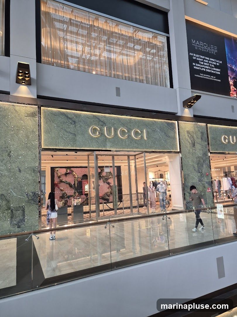 The exterior of a Gucci store with its name prominently displayed in illuminated letters on a green marble facade. The store has large glass windows and doors, revealing mannequins and merchandise inside. People are seen walking past the store.
