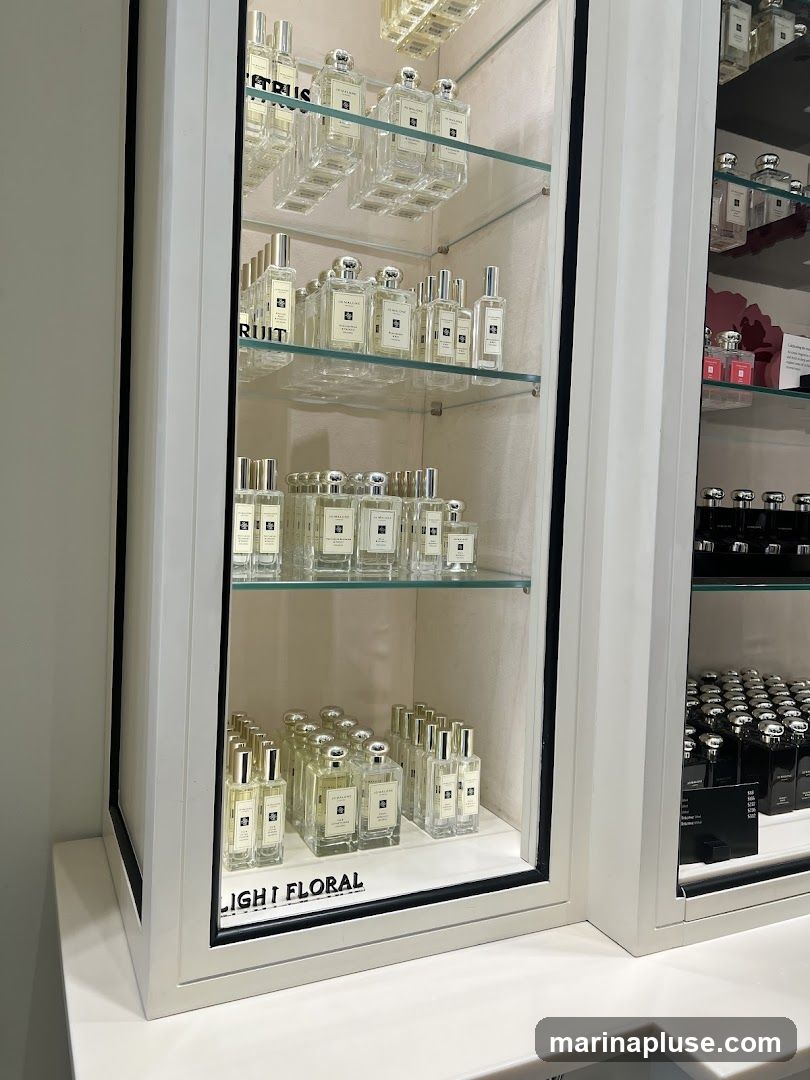 A display case filled with bottles of perfume. The bottles are arranged on glass shelves within a white cabinet. Some shelves have labels indicating scent categories like "CITRUS" and "LIGHT FLORAL". On the right side of the image, another display case shows rows of black perfume bottles.
