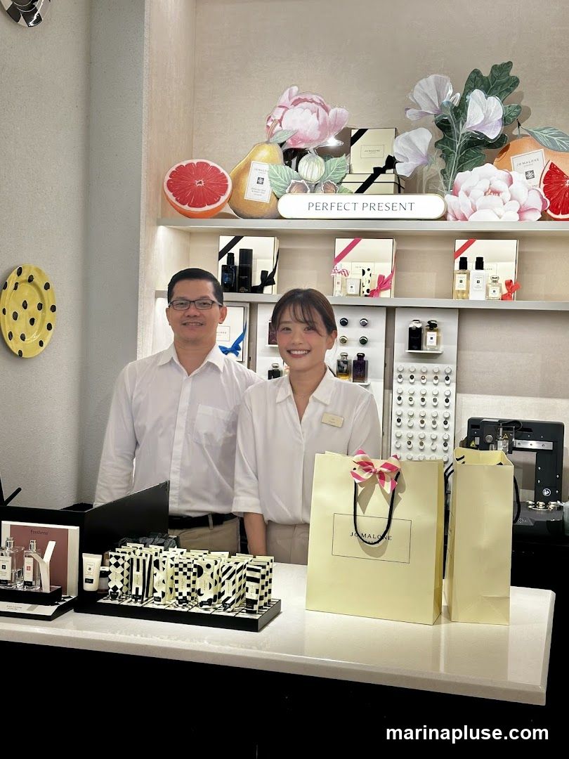 Two staff members stand behind a counter in a brightly lit store. The shelves behind them are stocked with various perfume bottles and gift sets. A decorative display features fruit illustrations and the words "PERFECT PRESENT." In the foreground, there are perfume bottles, a display of smaller items, and branded shopping bags.