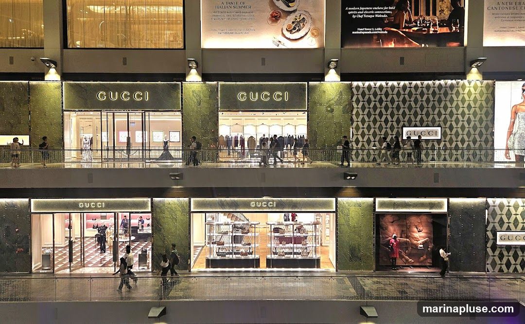 Multiple Gucci storefronts are visible on two levels of a shopping mall. The upper level features three distinct entrances, each with prominent "GUCCI" signage. The storefronts display mannequins and clothing racks. The lower level also shows multiple Gucci entrances, with displays of handbags and other merchandise. People are seen walking on the balconies in front of the stores. Above the upper level, large advertisements are displayed.