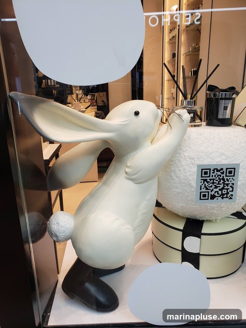 A window display features a large white rabbit sculpture with black boots, positioned next to a round, textured white object and a gift box. Behind these items, shelves are visible displaying various products, including diffusers and candles, with the word "SEPHORA" reflected in the glass. A QR code is also present on the display.