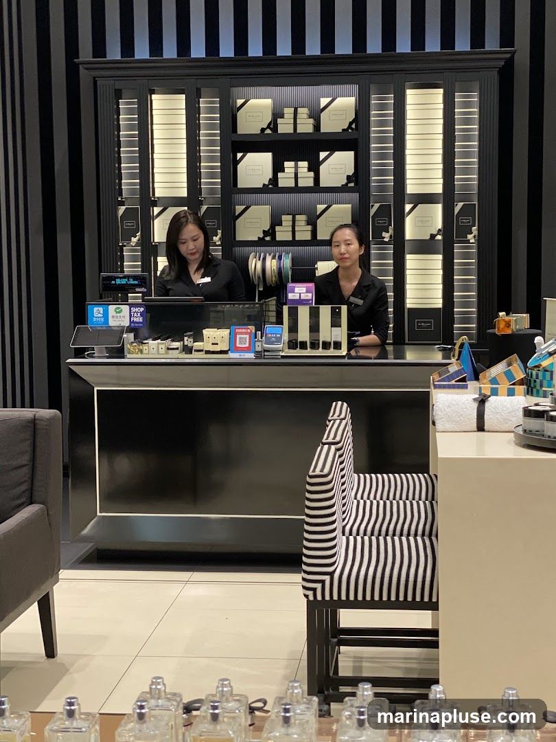 Two women stand behind a counter in a perfume store. The counter is black with a white trim. Behind them, shelves are stocked with boxes and bottles, many of which are branded with the Jo Malone logo. There are also display shelves with lighting. In the foreground, rows of perfume bottles are visible.