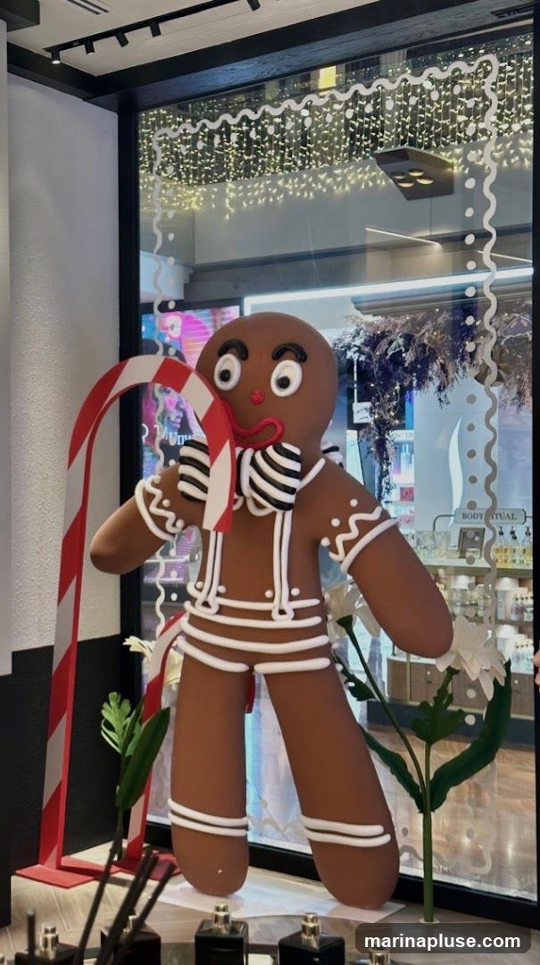 A large gingerbread man decoration is displayed in a shop window, holding a candy cane. The window is decorated with white wavy lines and fairy lights. In the background, shelves with bottles and products are visible, along with a sign that reads "BODY RITUAL".