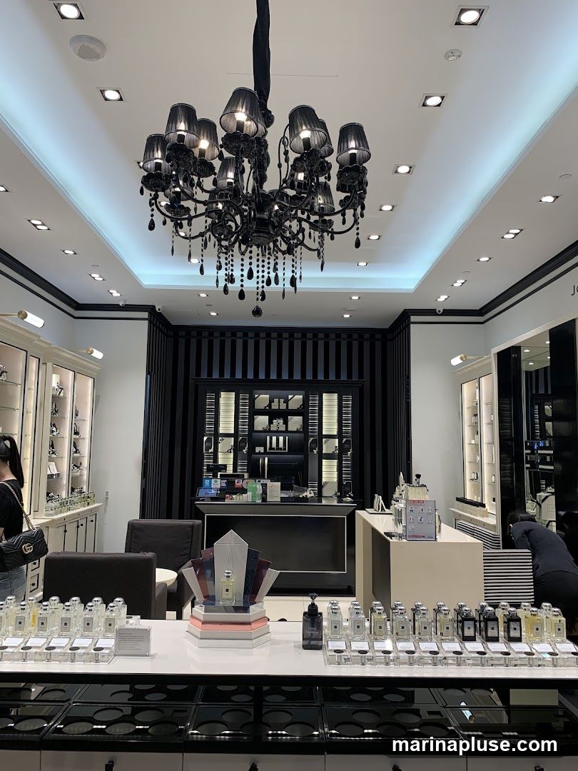 The interior of a perfume store with a large black chandelier hanging from the ceiling. Display shelves line the walls, showcasing various perfume bottles and products. A central counter area features more perfume bottles arranged on a display, with a decorative stand holding a prominent perfume bottle.
