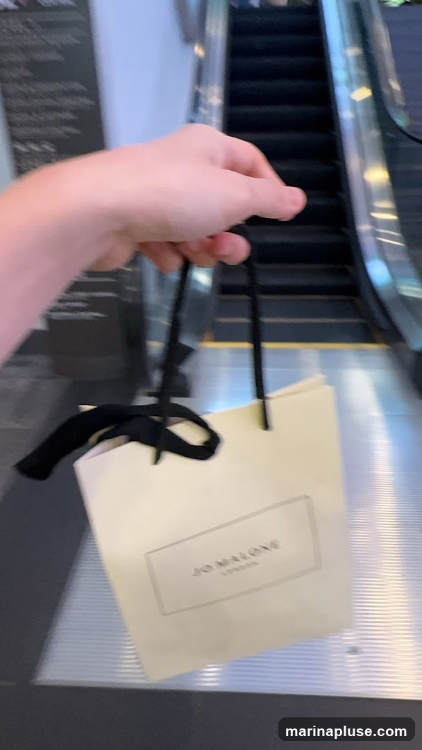 A hand holds a light-colored shopping bag with black handles and a black ribbon tied to one handle. The bag has a rectangular logo with text that appears to read "JO MALONE LONDON". The bag is positioned in front of a blurred escalator.