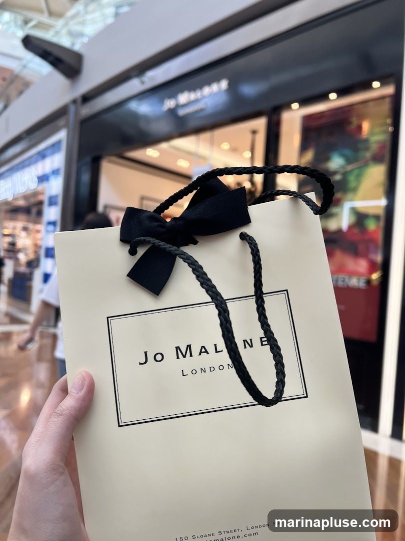 A hand holds a cream-colored Jo Malone shopping bag with black braided handles and a black bow. The Jo Malone London logo is printed on the front of the bag. In the background, the storefront of a Jo Malone store is visible, with its name illuminated above the entrance.