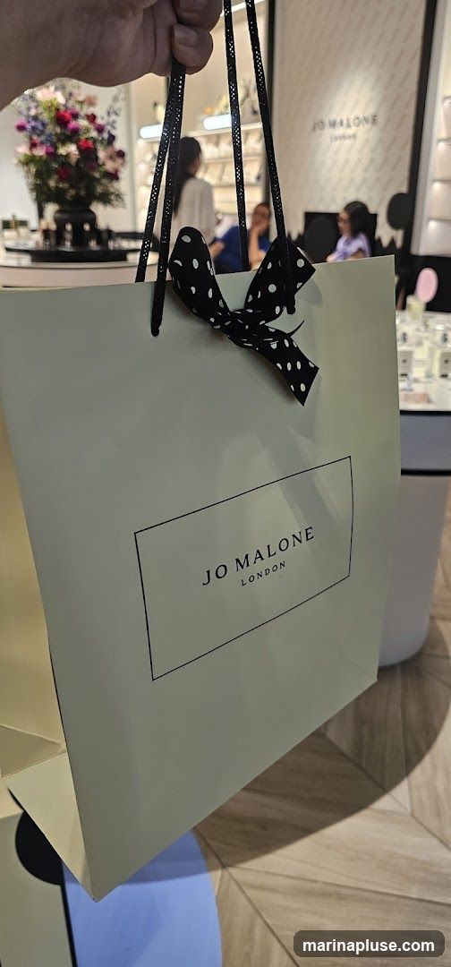 A light green shopping bag with the Jo Malone London logo printed in black on a white rectangle. The bag has black handles and a black and white polka dot ribbon tied in a bow. In the background, the interior of a store is visible, with shelves displaying products and people browsing.