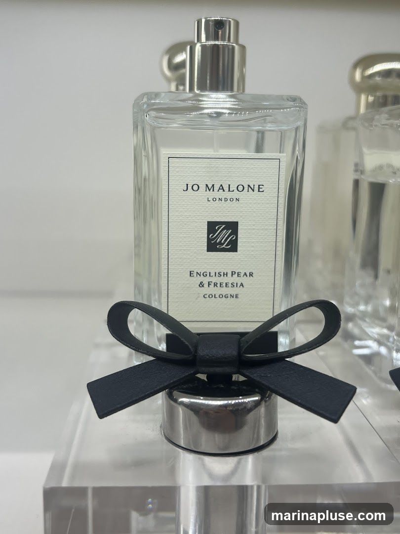 A close-up shot of a Jo Malone London English Pear & Freesia Cologne bottle. The clear glass bottle has a white label with black text and a black ribbon tied around the silver cap. Another bottle is visible in the background.