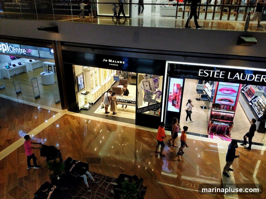 An overhead view of a shopping mall interior, showcasing the storefronts of several cosmetic and electronics retailers. Visible brands include Jo Malone London, Estée Lauder, and Epicentre. People are seen walking through the mall and browsing the stores. The floors are highly polished, reflecting the lights and displays.