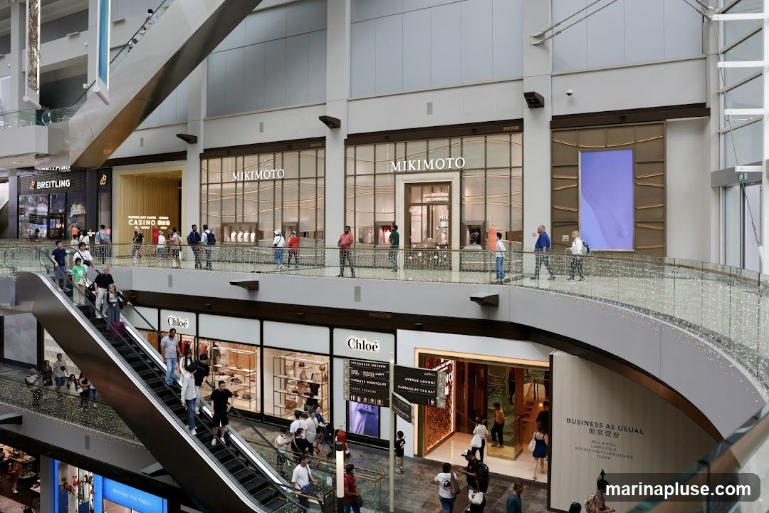 A view of a multi-level shopping mall with various luxury stores and escalators. Visible stores include Breitling, Mikimoto, and Chloe. People are seen shopping and using the escalators.