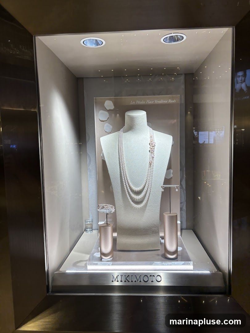 A well-lit display window showcases a pearl necklace and bracelet on mannequins. The brand name 'MIKIMOTO' is visible on the bottom of the display. The background features a subtle floral motif and the text 'Les Pétales Place Vendôme Rosés'.