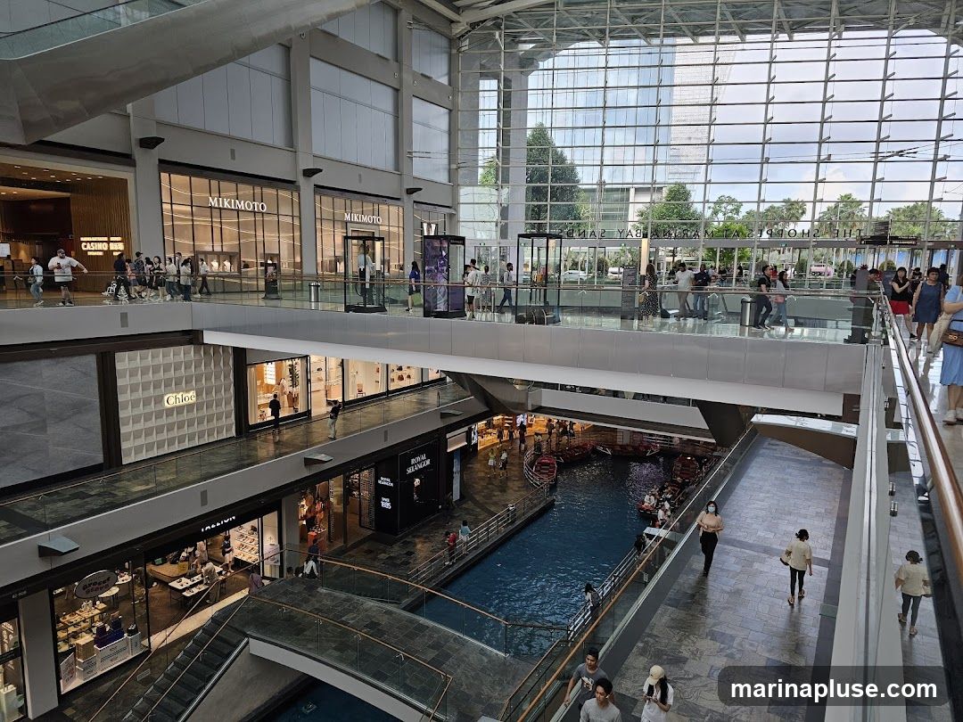 The image shows the interior of a modern, multi-level shopping mall. A canal with gondola-like boats is visible on a lower level. Shops with prominent signage like MIKIMOTO and Chloe are seen on the upper levels. People are walking on the elevated walkways and escalators. The mall features a large glass ceiling and walls, allowing natural light to enter.