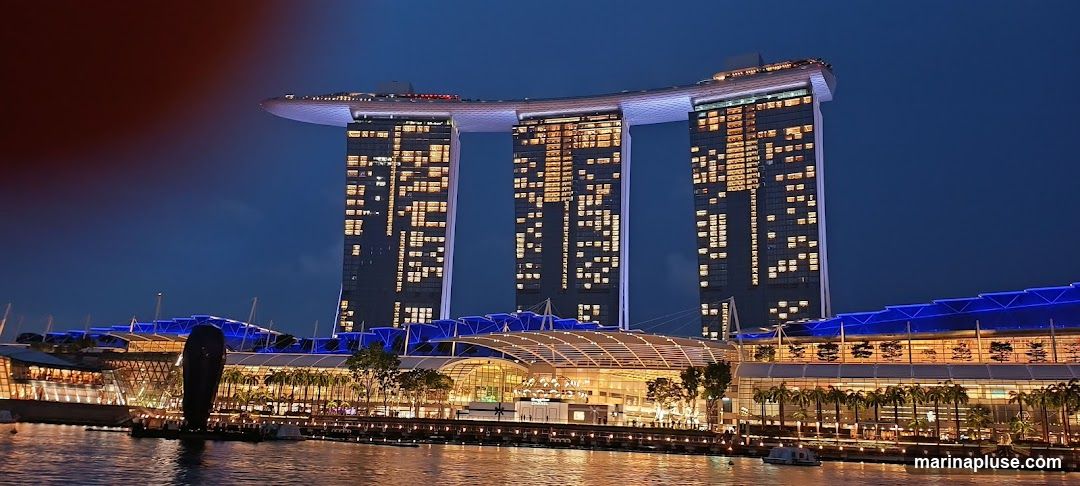 A wide shot of the Marina Bay Sands hotel and complex in Singapore at night. The iconic three towers are illuminated, with lights visible in many windows. The lower levels of the complex, featuring a modern architectural design with blue lighting accents, are also visible, as is the waterfront promenade.