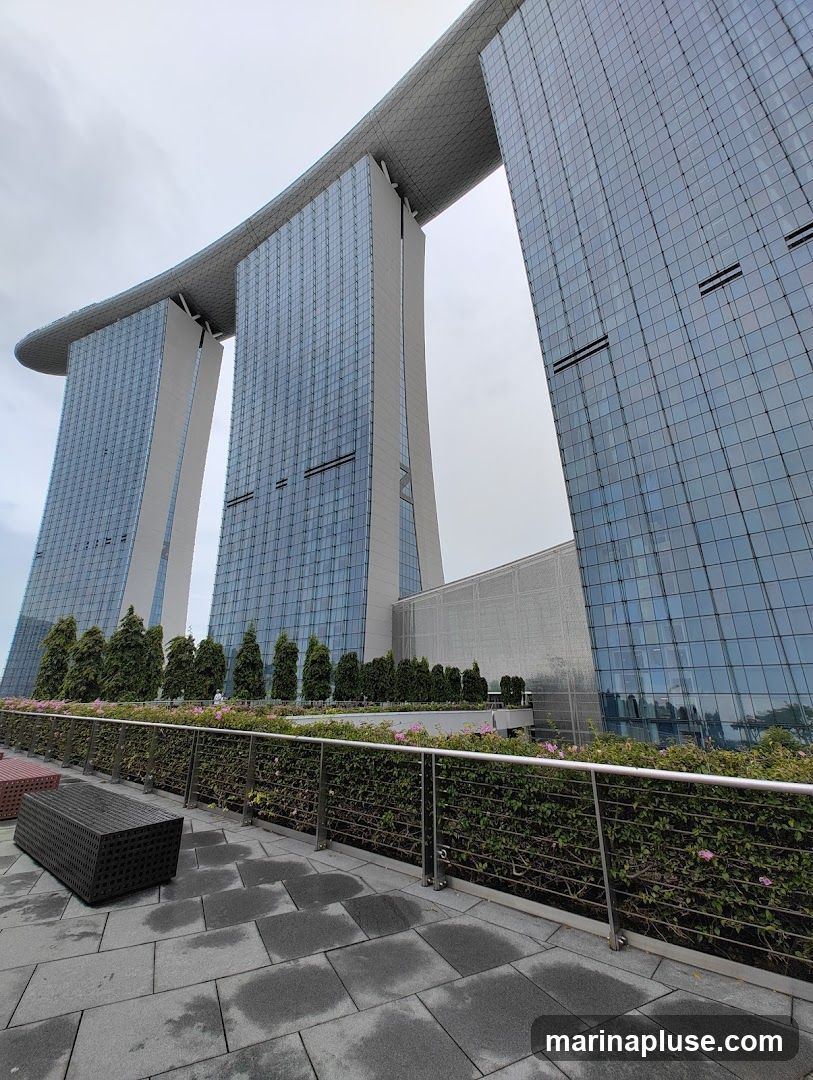A low-angle shot of the Marina Bay Sands hotel in Singapore on an overcast day. The three towers are connected by a large, curved structure at the top. In the foreground, there is a paved walkway with a low hedge and railing. A dark, woven bench is visible on the left.
