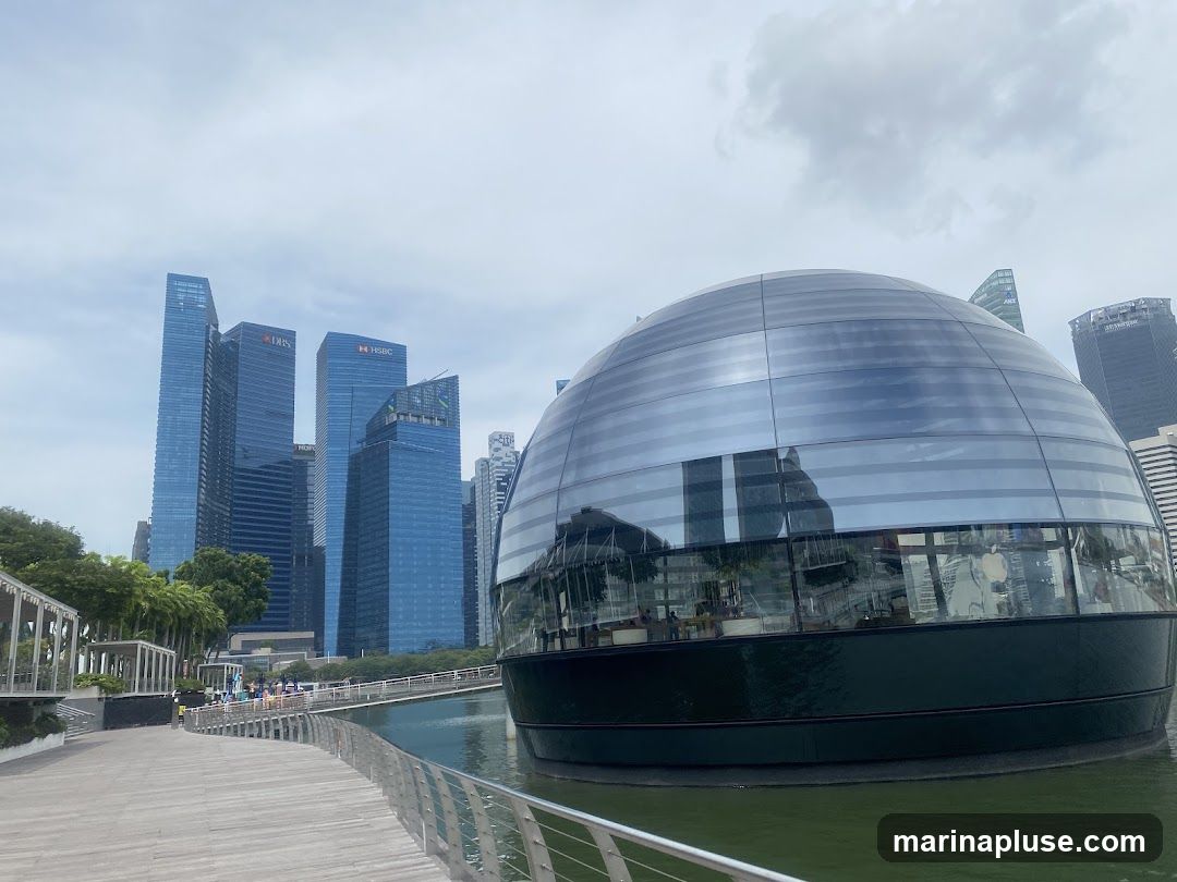 A wide shot of a futuristic spherical building with a glass facade, situated on the water. In the background, several tall, modern skyscrapers dominate the skyline under a cloudy sky. A wooden walkway with a metal railing curves along the foreground, leading towards the spherical building. People can be seen walking on a bridge in the distance. The reflection of the Apple logo is visible on the glass facade of the spherical building.
