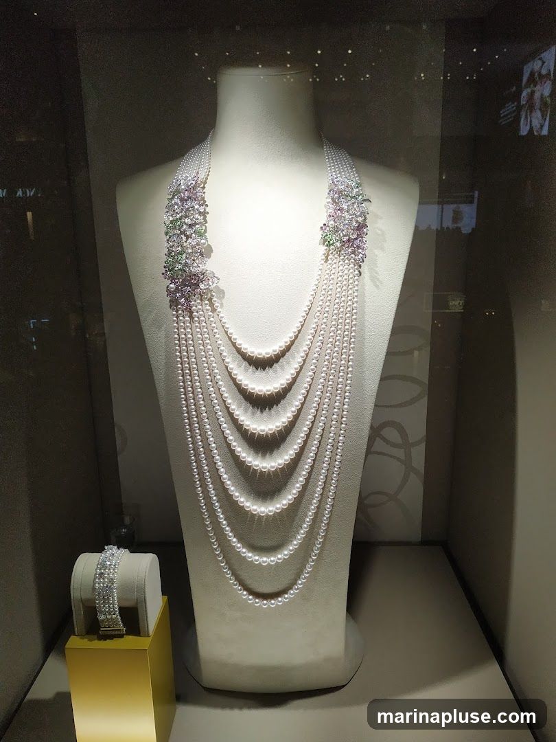 A multi-strand pearl necklace with floral gemstone accents is displayed on a mannequin bust. A diamond bracelet is showcased on a gold stand to the left. The display is set within a dimly lit, elegant jewelry store window.