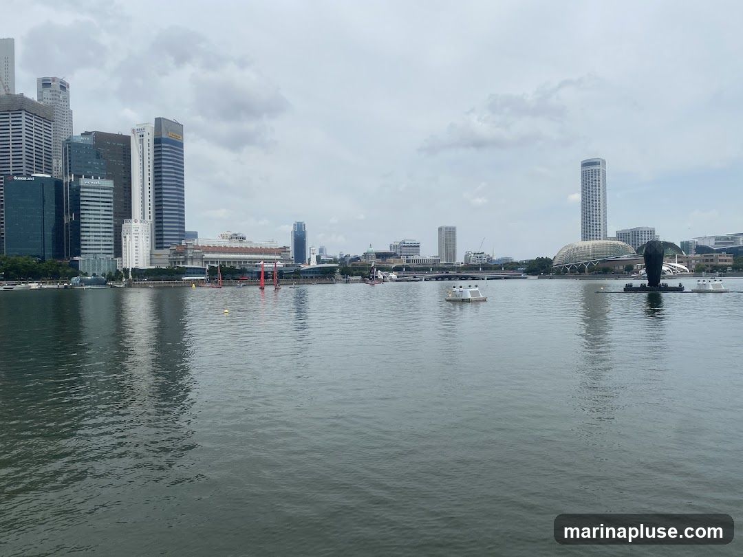 A wide shot of the Marina Bay skyline in Singapore on an overcast day. The water in the foreground reflects the tall buildings and cloudy sky. Several boats and floating structures are visible on the bay. In the distance, iconic buildings like the Marina Bay Sands and the ArtScience Museum can be seen, along with numerous skyscrapers.