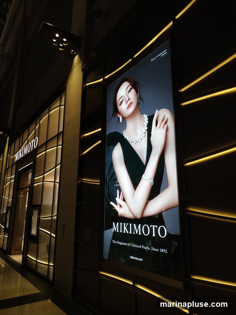 A large illuminated advertisement for Mikimoto features a woman wearing pearl jewelry. The advertisement displays the Mikimoto logo and the tagline "The Originator of Cultured Pearls. Since 1893." The store facade with the Mikimoto signage is visible to the left.