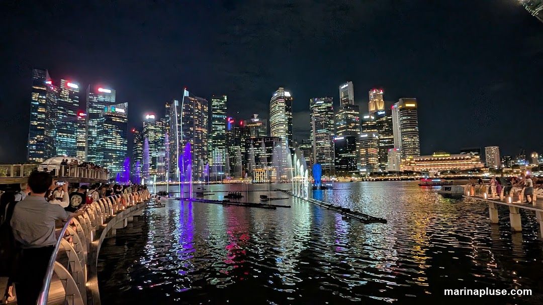 A nighttime view of a modern cityscape with numerous illuminated skyscrapers. In the foreground, a body of water reflects the city lights and a colorful light and water show. A curved walkway with people watching the show is visible on the left, and a pier with more spectators is on the right.