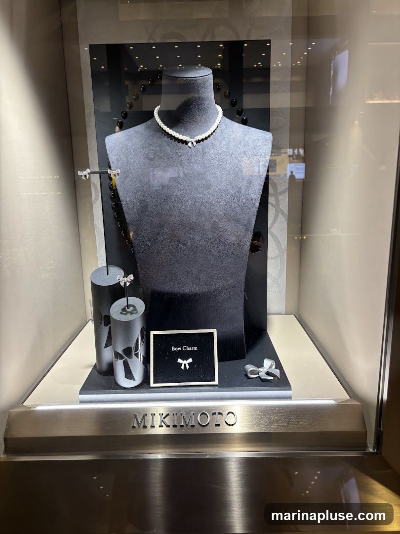 A display case featuring a pearl necklace with a bow charm on a mannequin bust. To the left, earrings and cylindrical stands with bow designs are visible. A small sign reads "Bow Charm" with a bow icon. A silver bow charm rests on the right side of the display. The brand name "MIKIMOTO" is visible on the front of the display case.