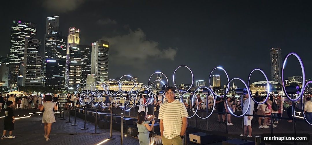 A panoramic view of a city skyline at night, with illuminated skyscrapers in the background. In the foreground, a man stands amidst a display of large, glowing rings that form an abstract sculpture. Several people are visible, some interacting with the installation, while others stroll along a wooden promenade. The overall scene suggests an outdoor event or exhibition in an urban setting.