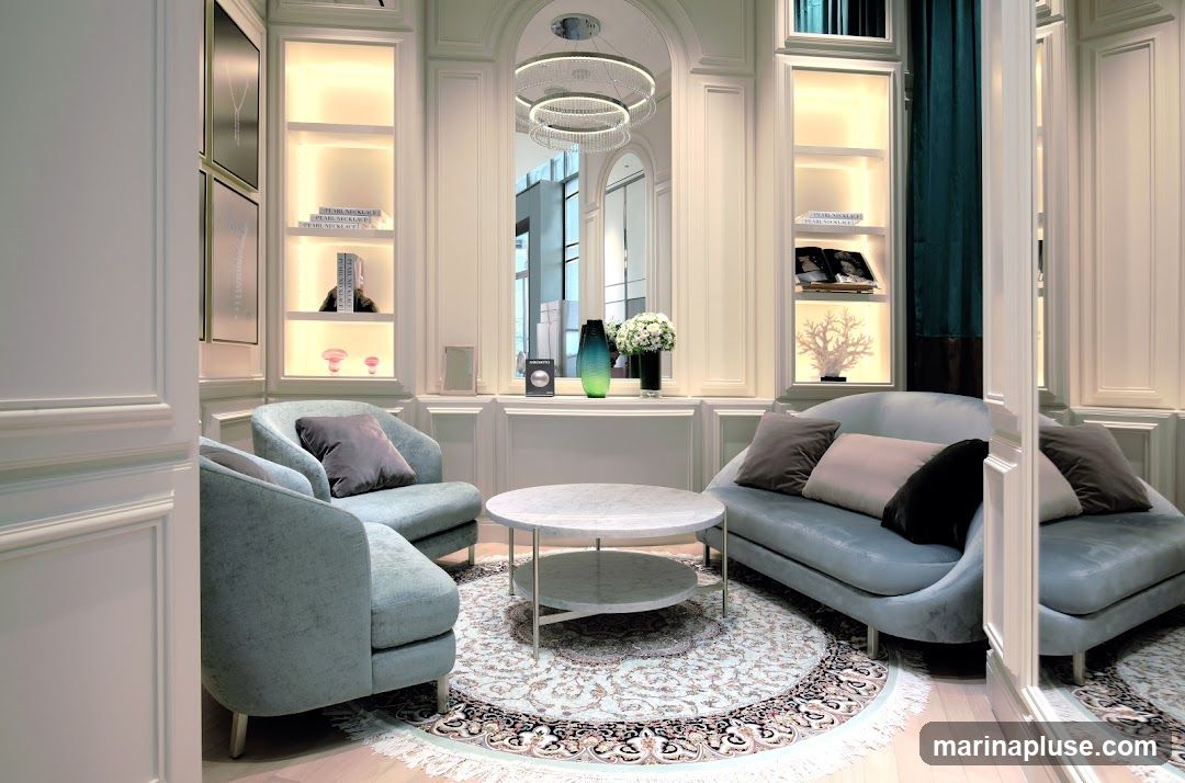 A luxurious and elegantly designed sitting area featuring plush light blue sofas and armchairs arranged around a marble coffee table. The room is adorned with decorative shelves displaying books, vases, and coral sculptures, illuminated by soft, warm lighting. A large mirror reflects the interior, enhancing the sense of space and sophistication. A patterned rug anchors the seating arrangement, adding a touch of classic charm.
