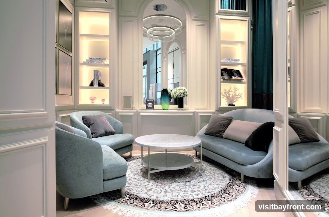 A luxurious and elegantly designed sitting area featuring plush light blue sofas and armchairs arranged around a marble coffee table. The room is adorned with decorative shelves displaying books, vases, and coral sculptures, illuminated by soft, warm lighting. A large mirror reflects the interior, enhancing the sense of space and sophistication. A patterned rug anchors the seating arrangement, adding a touch of classic charm.