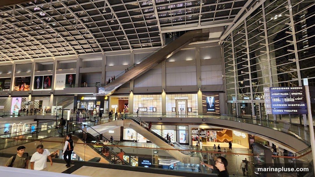 The image shows the interior of a modern shopping mall with multiple levels, escalators, and various shops. The architecture features a high, glass-paneled ceiling and a prominent, angled escalator. Signs indicate various destinations such as ArtScience Museum, Event Plaza, Spectra, Food Court, Digital Light, Marquee Nightclub, and Sands Theatre. Shops visible include Dior, Breitling, Mikimoto, and Royal Selangor. People are seen walking around and using the escalators.