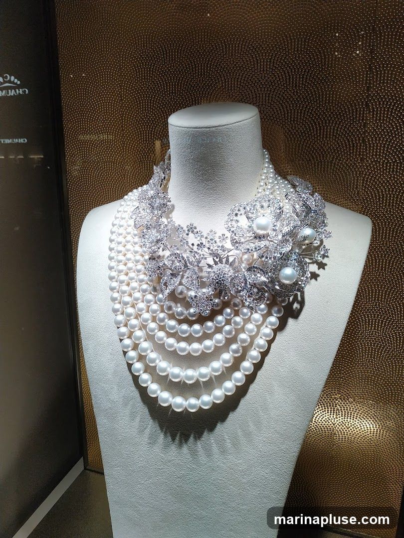 A multi-strand pearl necklace with a diamond and pearl encrusted floral brooch is displayed on a white mannequin bust. The background is a textured gold wall. A reflection of a store sign is visible on the left side of the image.