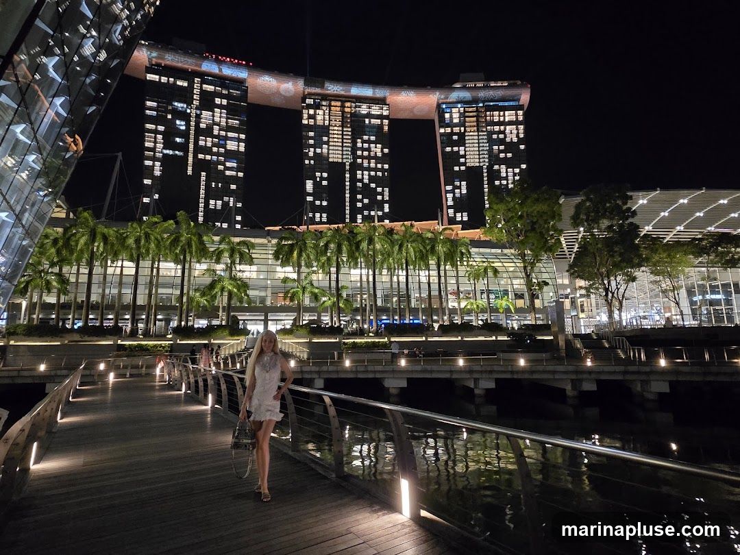 A woman in a white dress walks on a wooden boardwalk at night. She is carrying a handbag. In the background, a large, modern building complex is illuminated with many lights. Palm trees line the front of the building. The building's facade is decorated with illuminated patterns. The boardwalk has metal railings and is lit by embedded lights. The water reflects the lights from the boardwalk and the building.