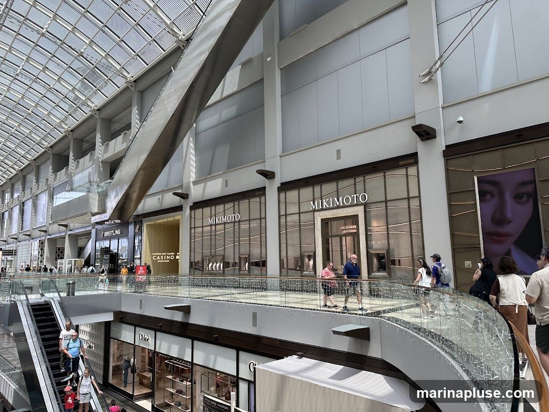 The image shows the exterior of a Mikimoto store within a modern shopping mall. The store has a large glass facade with the Mikimoto logo prominently displayed above the entrance. Inside, display cases with jewelry are visible. Other stores, including Breitling and Chloe, are also visible in the background, along with escalators and shoppers. A large advertisement featuring a woman's face is to the right of the Mikimoto store.