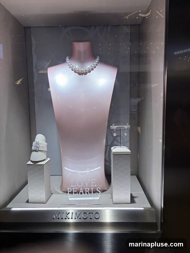 A display case featuring a pearl necklace on a mannequin bust, a pearl bracelet on a stand to the left, and pearl earrings on a stand to the right. A circular sign in front of the mannequin reads "LOVE PEARLS". The brand name "MIKIMOTO" is visible at the bottom of the display.