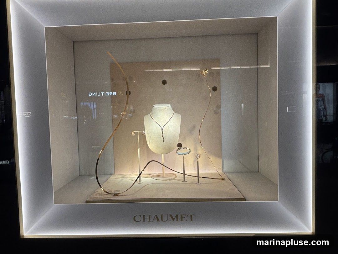 A well-lit jewelry display window featuring a necklace and bracelet on mannequins. The brand name "CHAUMET" is visible on the bottom frame of the window. Reflections of "BREITLING" and "CASINO" are visible in the glass.