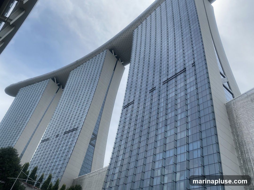 A low-angle shot of the Marina Bay Sands hotel in Singapore, showcasing its distinctive architecture with three towers connected by a large, curved rooftop structure. The building is clad in glass and steel, reflecting the cloudy sky.