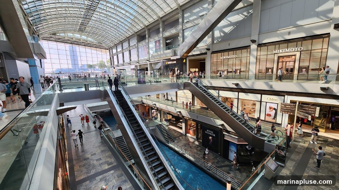 An interior view of a multi-level shopping mall with escalators, shops, and a water feature. People are seen walking around and shopping. Shops visible include MIKIMOTO, Chloe, Breitling, and Royal Selangor. The mall has a large glass roof allowing natural light to enter.