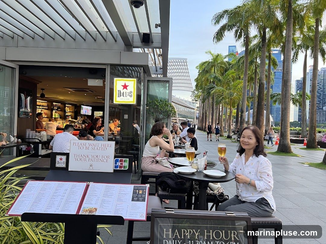 A woman sits at an outdoor table at Dallas Cafe & Bar, holding a glass of beer. In the background, palm trees line a walkway with a view of city skyscrapers. The cafe's entrance and bar area are visible, with patrons seated inside and outside. A menu stand is placed in front of the seating area.