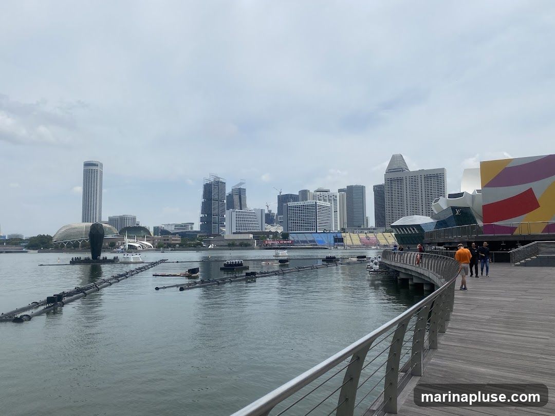 A wide wooden promenade curves along a body of water, with a city skyline in the background. Several modern buildings, including a tall skyscraper and a distinctive domed structure, are visible. In the foreground, floating structures with pipes are present in the water. People are walking on the promenade. The building on the right features a colorful, abstract design.