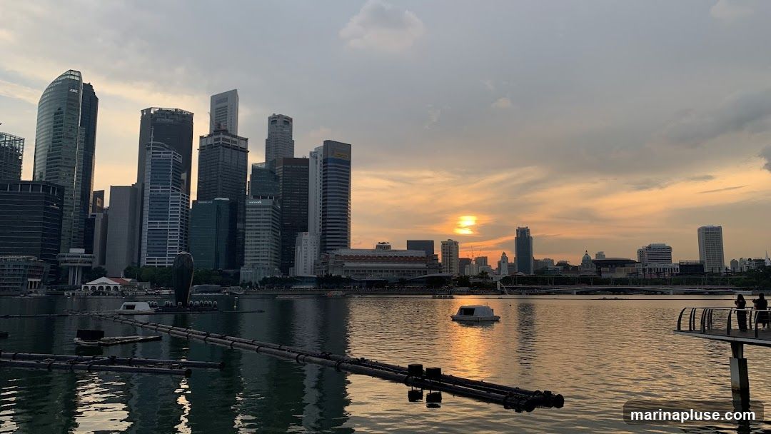 A panoramic view of a modern cityscape with numerous skyscrapers reflected in the water. The sun is setting, casting a warm orange glow across the sky and water. Several boats and floating structures are visible on the water. In the foreground, there are some floating pipes or booms. On the right, a pier with two silhouetted figures overlooks the scene.