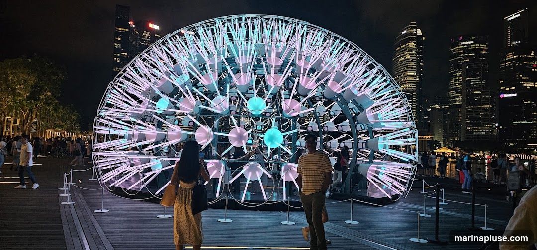 A large, spherical light installation with numerous glowing tubes and orbs is displayed outdoors at night. The installation is illuminated with pink and blue lights. In the background, a city skyline with illuminated skyscrapers is visible. People are walking around the installation and along a wooden boardwalk.