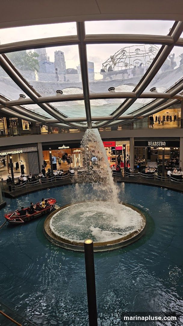 An indoor shopping mall features a large circular water fountain with a powerful stream of water cascading down. A small boat with several passengers navigates the water. Surrounding the fountain are multiple shops and restaurants with patrons seated at tables. The mall has a modern design with a glass ceiling allowing natural light to filter in, and offers views of the city skyline outside. Visible shop names include Emporio Armani, Stone Island, Hugo, and Beanstro.