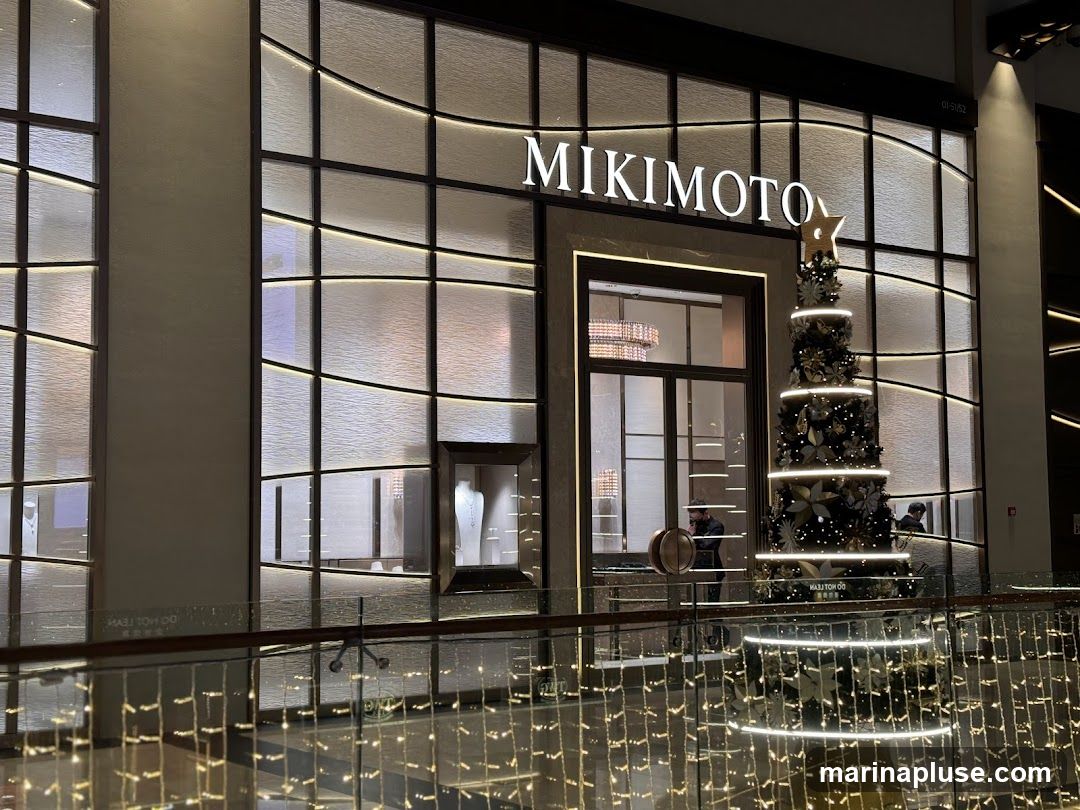 The exterior of a Mikimoto store is shown, featuring a large display window and the Mikimoto logo prominently displayed above the entrance. A decorated Christmas tree stands to the right of the entrance, illuminated with lights. The store's facade is modern and elegant, with textured glass panels and subtle lighting.