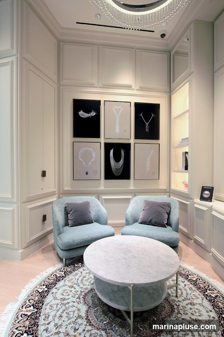 A luxurious jewelry store interior featuring a seating area with two light blue armchairs and a marble coffee table. The walls are adorned with framed images of various necklaces, some on black backgrounds and others on lighter backgrounds. One image prominently displays a necklace with a large blue sapphire. To the right, a lit display shelf holds books and a small framed item. A circular rug with intricate patterns lies beneath the coffee table. A modern chandelier hangs from the ceiling.