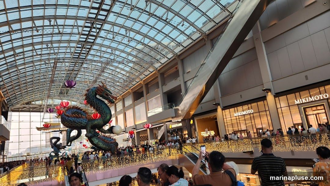An indoor mall with a large, ornate dragon decoration suspended from the ceiling. The dragon is green and blue with red and purple accents. It is surrounded by festive lights and lanterns. Many people are visible on multiple levels of the mall, looking at the decoration and shopping. Storefronts with prominent signage, including