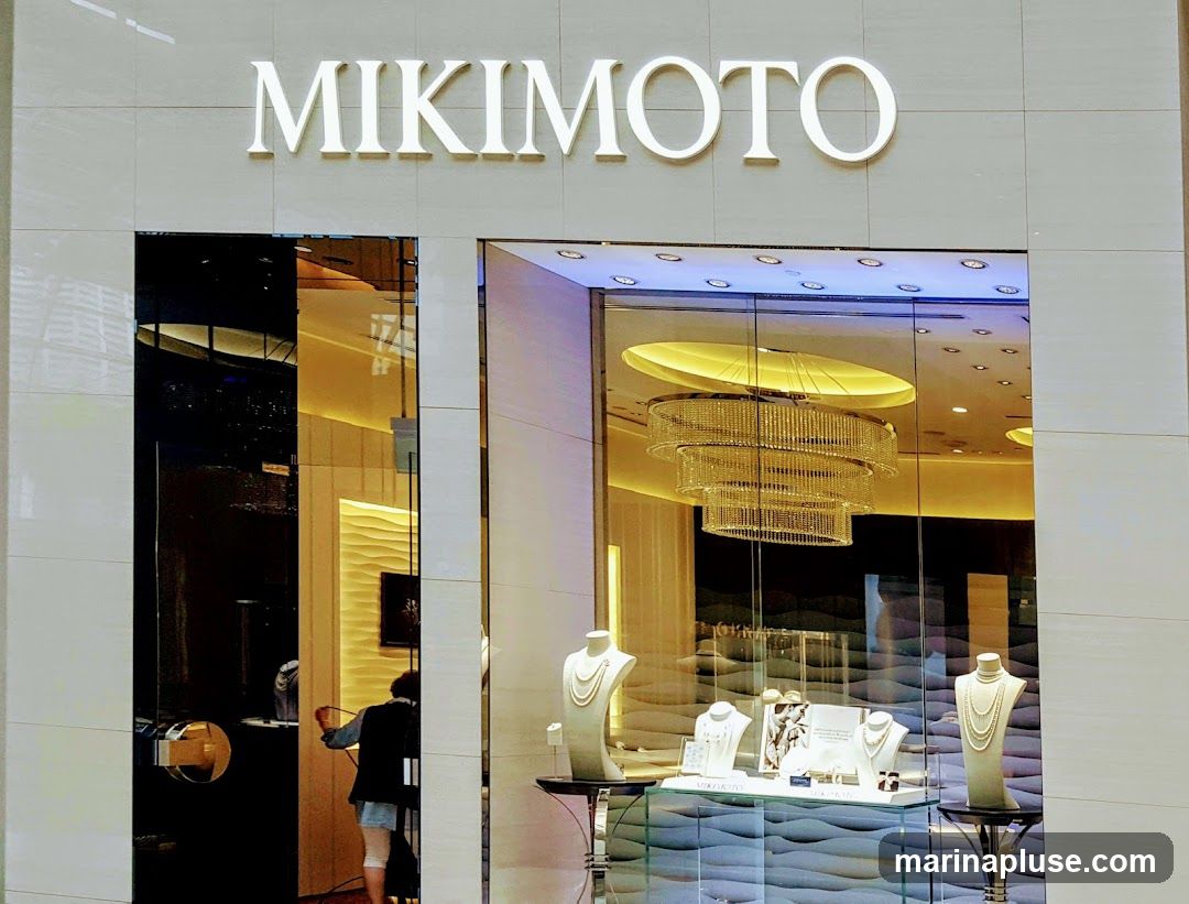 The exterior of a Mikimoto jewelry store. The store name is displayed in large white letters above a glass storefront. Inside, a chandelier hangs from the ceiling, and jewelry is displayed on mannequins and stands. A person is visible inside the store, near the entrance.