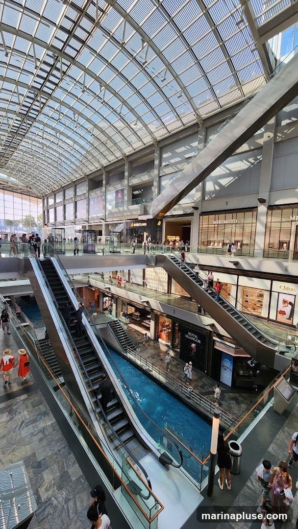 An interior view of a multi-level shopping mall with a glass ceiling. Escalators ascend between floors, and a canal with blue water runs through the center of the mall. Various shops are visible on different levels, including Breitling, Mikimoto, Chloe, and Royal Selangor. People are seen shopping and walking around.