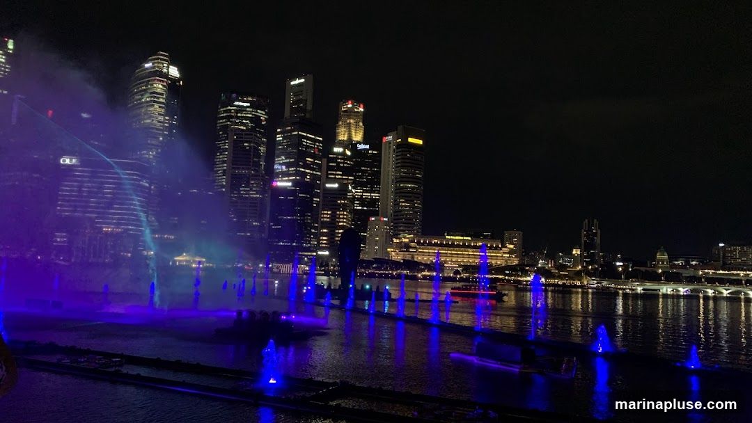 A nighttime view of a fountain show with colorful lights, set against a backdrop of a modern city skyline. The water jets are illuminated with blue and purple lights, creating a vibrant display on the water's surface. Tall skyscrapers with illuminated windows dominate the background, reflecting the city's energy. A boat is visible in the distance on the water.