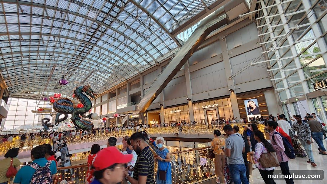 A large, ornate dragon decoration hangs from the ceiling of a spacious, modern mall. The mall features a glass roof, multiple levels, and numerous shops with illuminated signs. A large crowd of people are gathered on the walkways, observing the decoration and the shops. The overall atmosphere appears festive and busy.