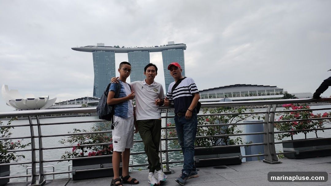 Three men are standing on a paved walkway, posing for a photo. Behind them, the iconic Marina Bay Sands hotel and the ArtScience Museum in Singapore are visible. The sky is overcast. The man on the left is wearing a blue and white tie-dye t-shirt, white shorts, and sandals. The man in the middle is wearing a white polo shirt with a logo, and dark green pants. The man on the right is wearing a red cap, a black and white striped t-shirt, and blue jeans. They are standing in front of a metal railing with some flowering plants.